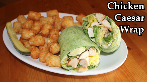A chicken caesar wrap in a spinach tortilla, halved and plated with tater tots and a pickle spear